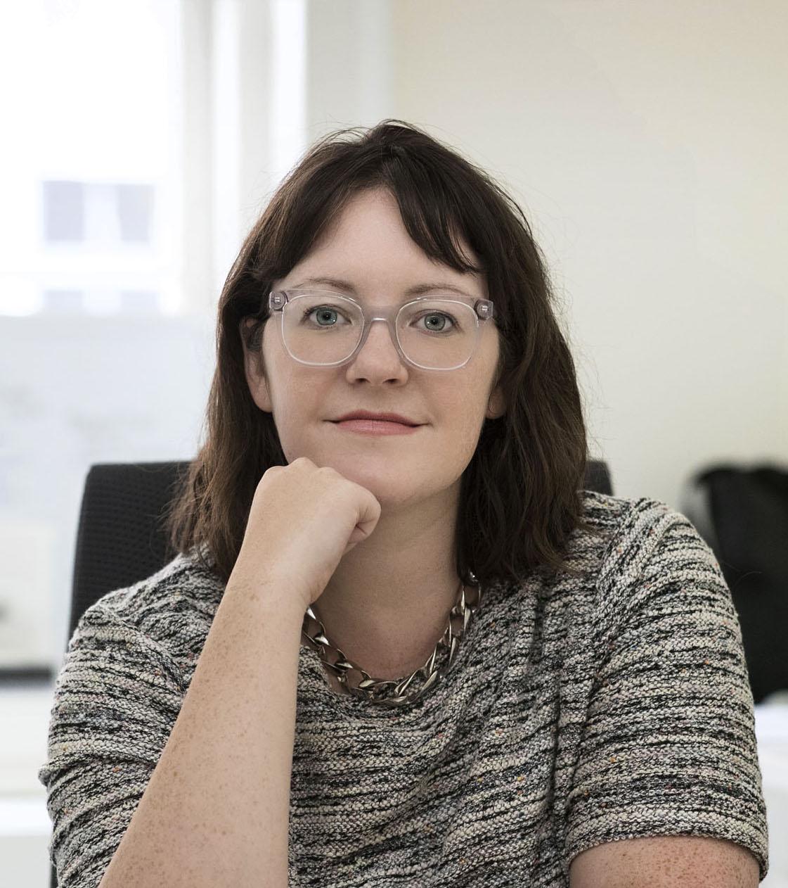 A white woman with dark hair, wearing clear glasses and a grey sweater