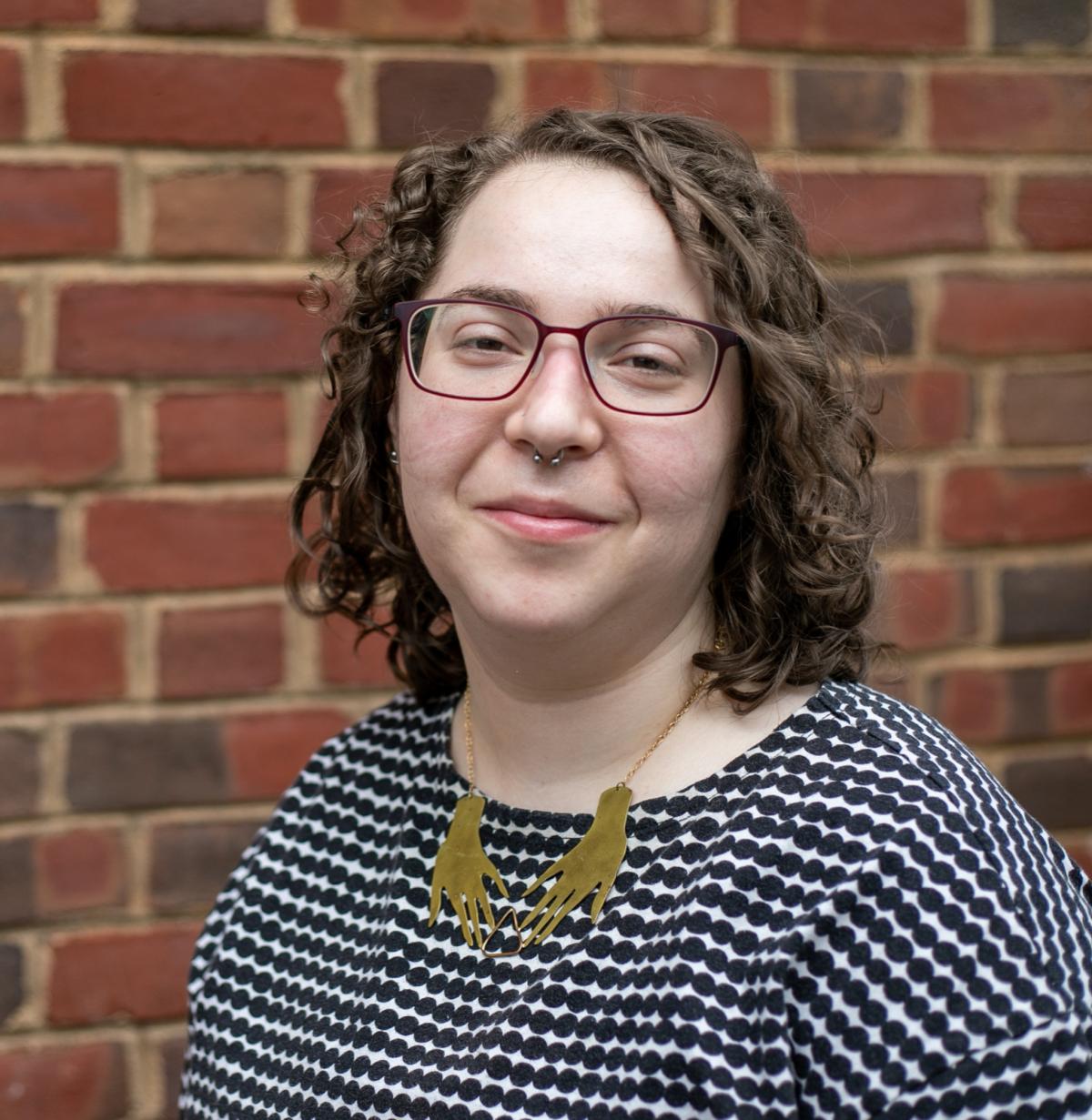 A white woman with curly hair and glasses in front of a brick wall